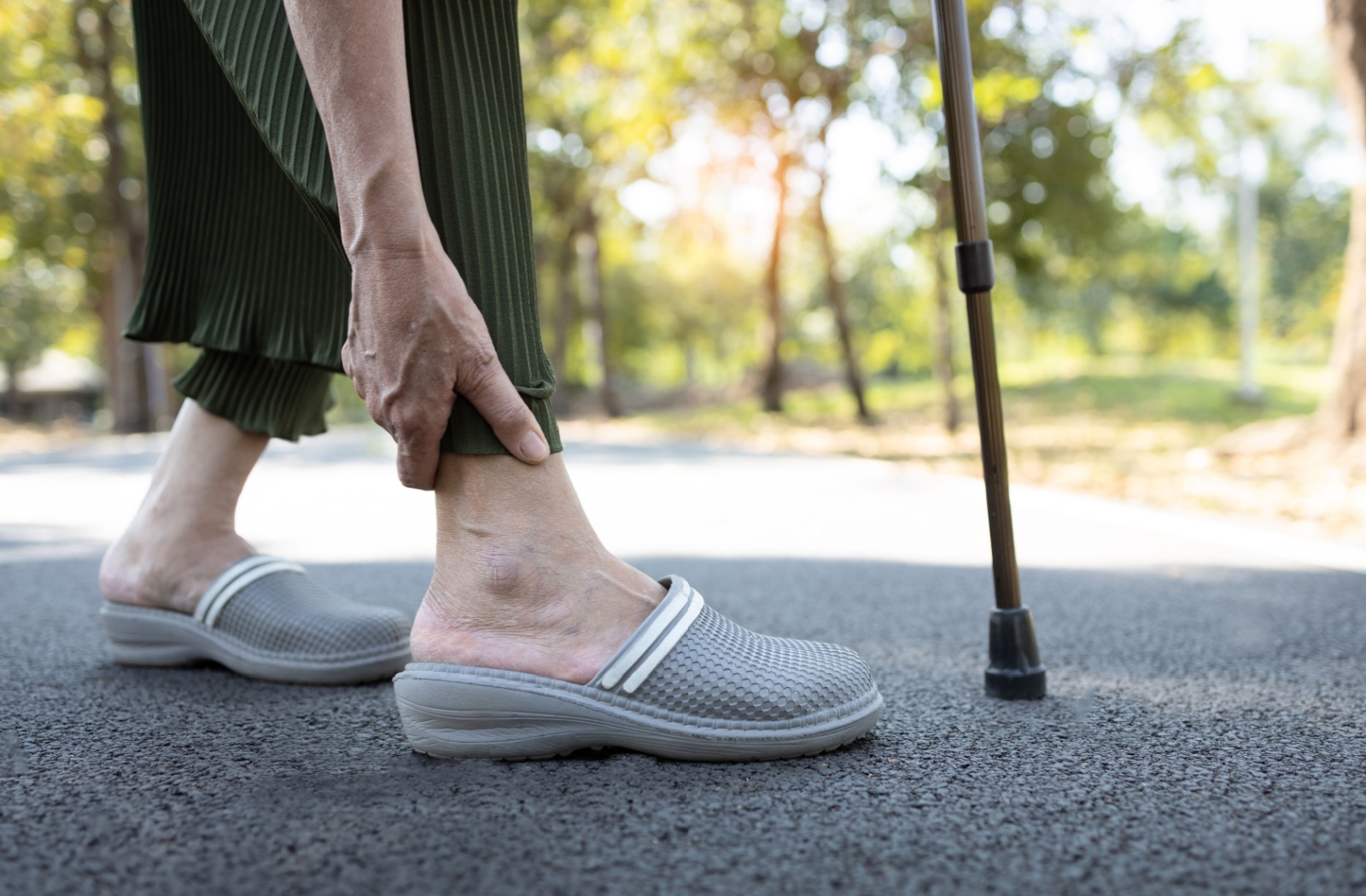 An older adult wearing slippers bending to rub their ankle while on a walk outside.