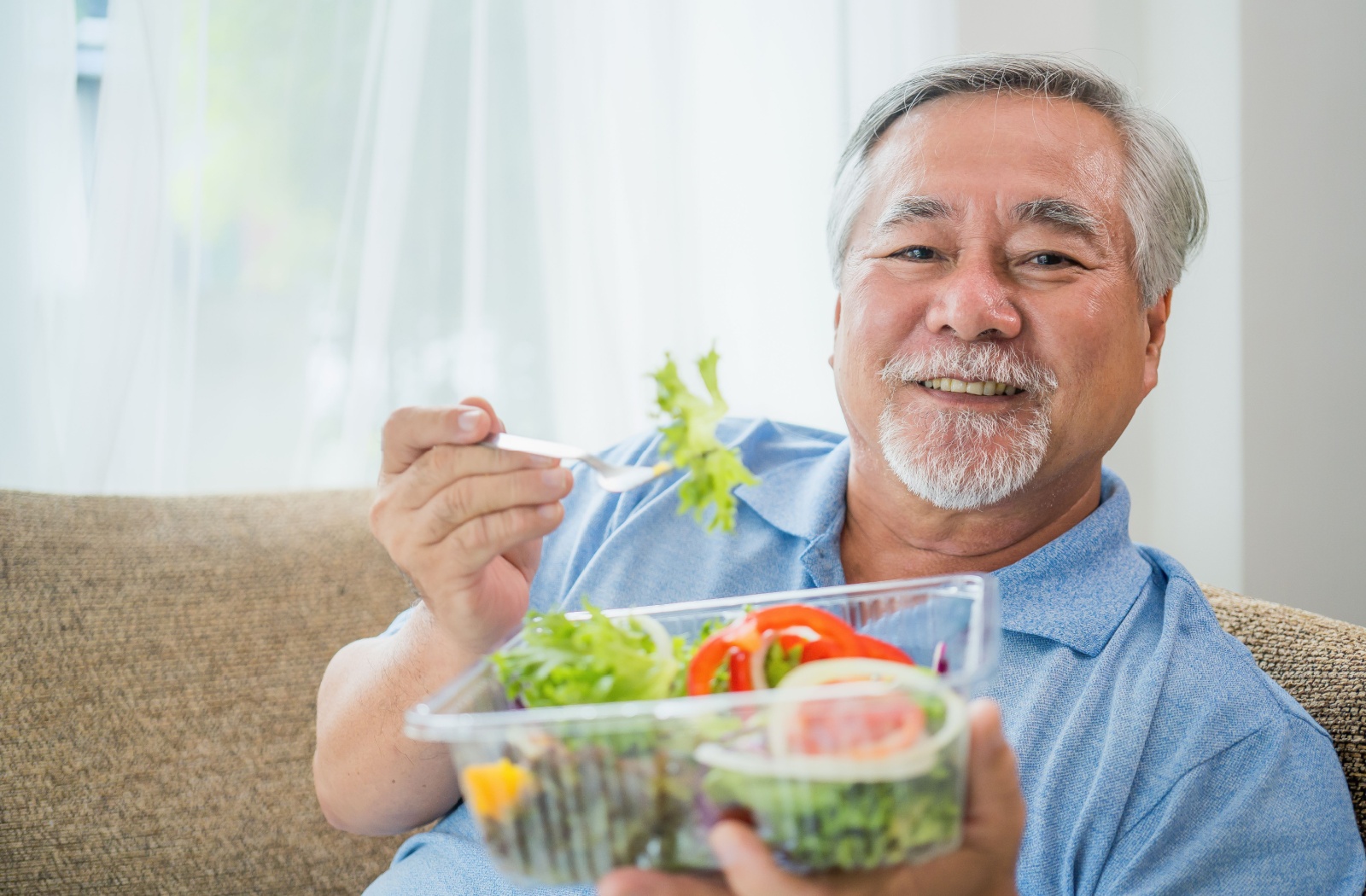 A senior smiles while holding a clear container of salad, enjoying a mix of leafy greens, tomato, and onion.