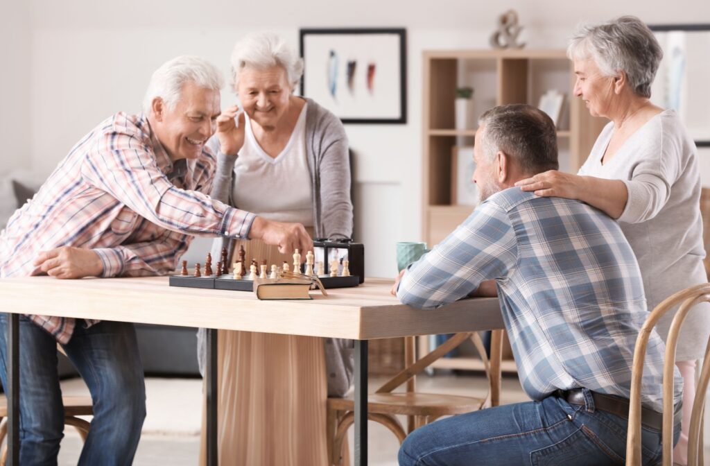 Two seniors play each other in chess in a communal area of their senior living community while others watch on